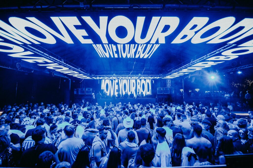 Indoor DJ set with a large crowd and neon blue lighting staying "MOVE YOUR BODY".