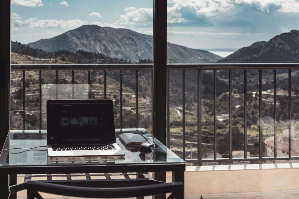 Remote work setup with laptop, mouse, and notebook on a table overlooking hills and water.
